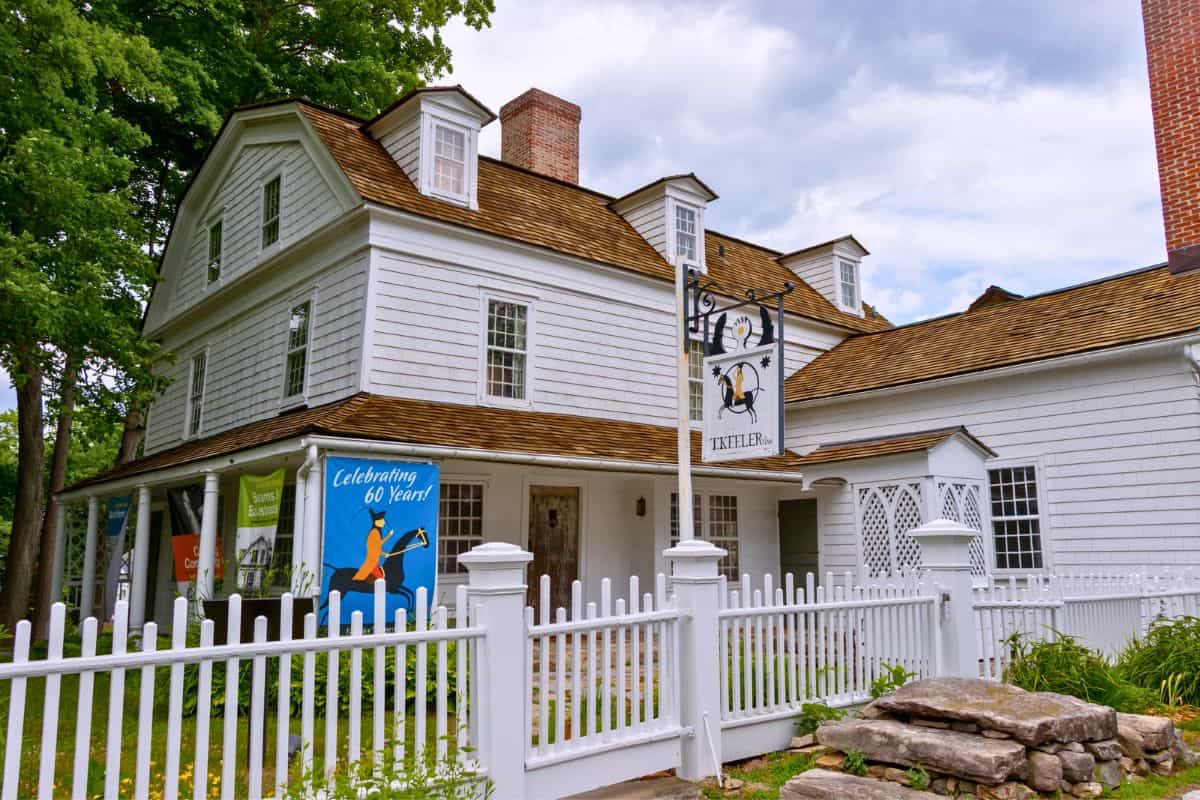 Two-story white colonial-style house with a gambrel roof, white picket fence, and a sign reading “Tavern.” A banner on the porch says “Celebrating 60 Years.”.
