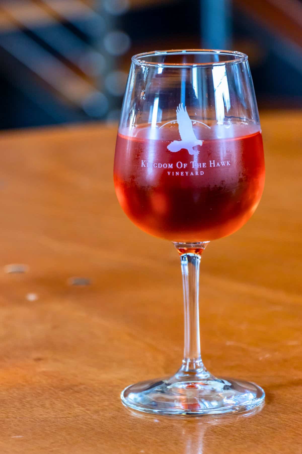 A glass of rosé wine on a wooden table, with "Kingdom Of The Hawk Vineyard" and a hawk logo printed on the glass.