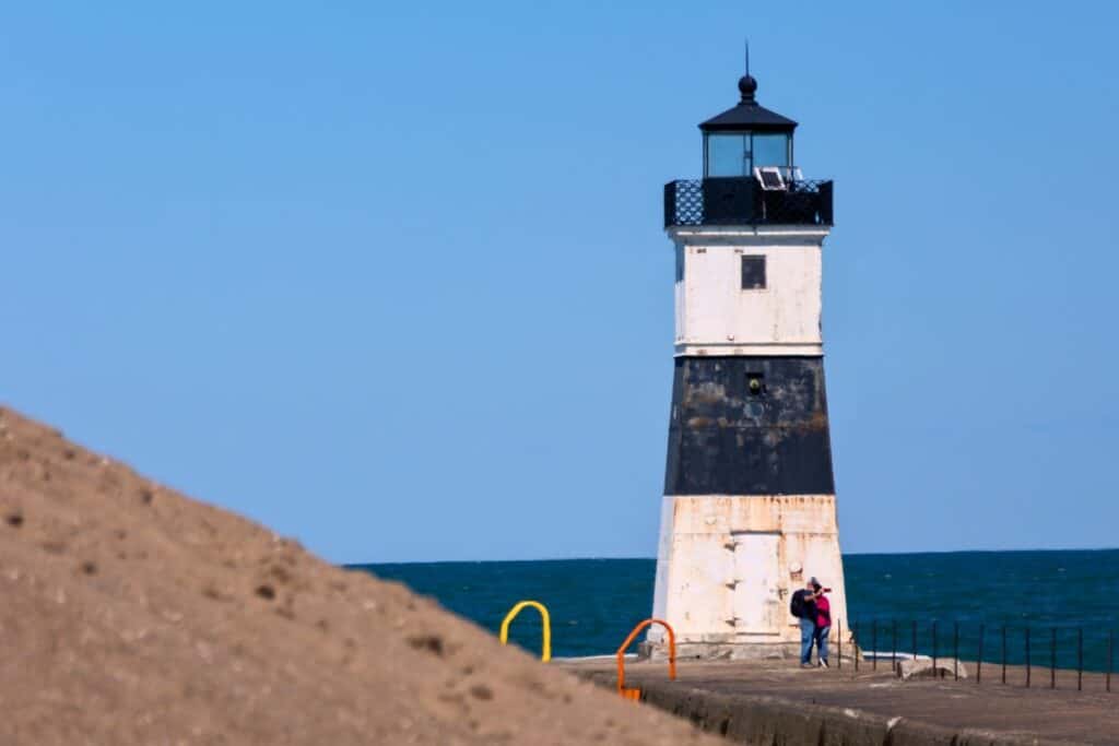 A white and black lighthouse stands on a pier by the ocean, with two people standing near its base under a clear blue sky.