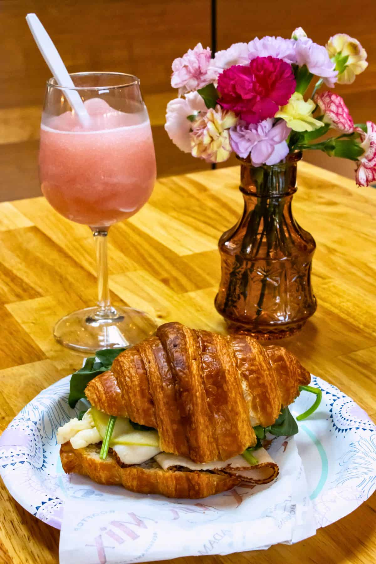 A croissant sandwich with greens and cheese on a paper plate, a glass of pink frozen drink with a straw, and a vase of colorful flowers on a wooden table.