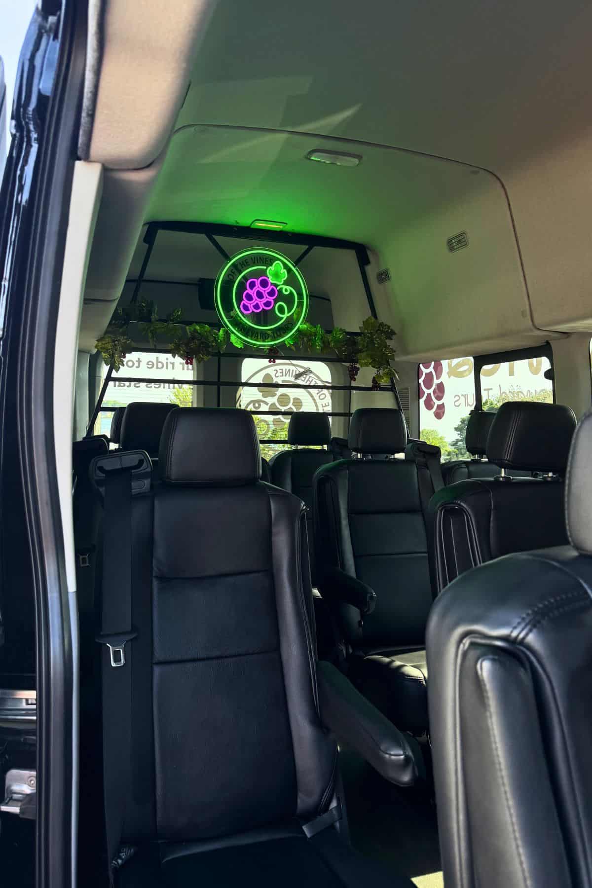 The interior of a passenger van with black leather seats, a neon grape sign, and grapevine decorations above the back window.