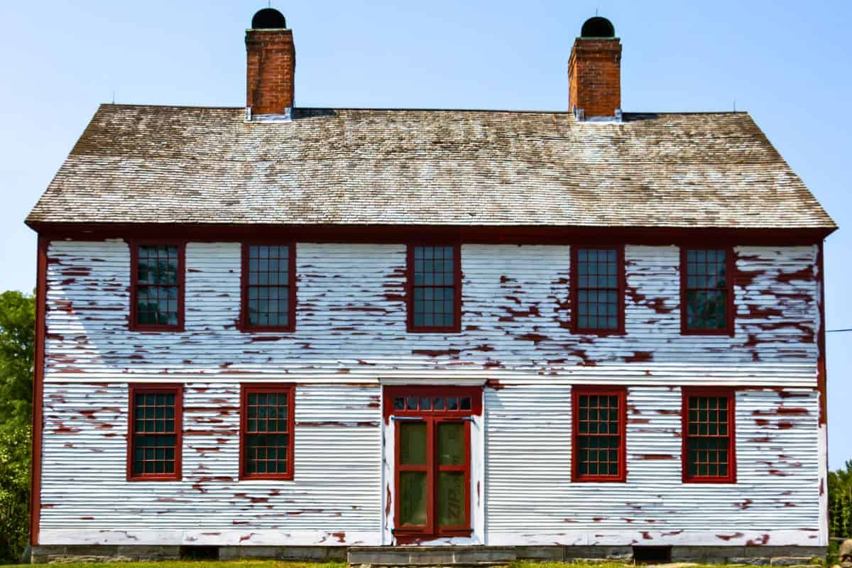 A two-story white wooden house with peeling paint, red trim, and two brick chimneys, viewed from the front against a clear sky.