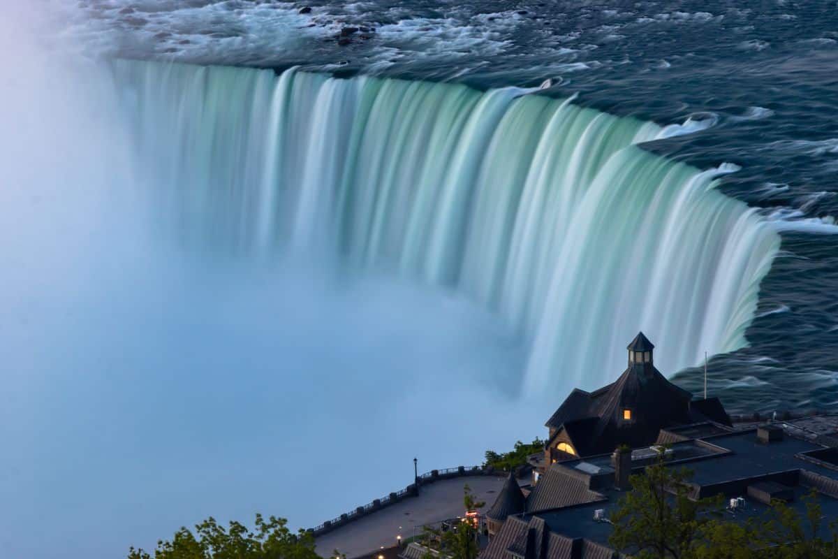 A large waterfall with water cascading over the edge, creating mist. A dark-roofed building is visible in the foreground.