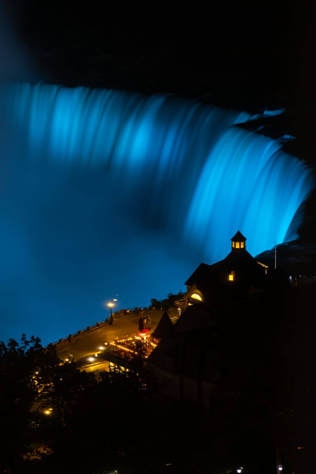 Niagara Falls illuminated in blue at night, with a building and street lights visible in the foreground.