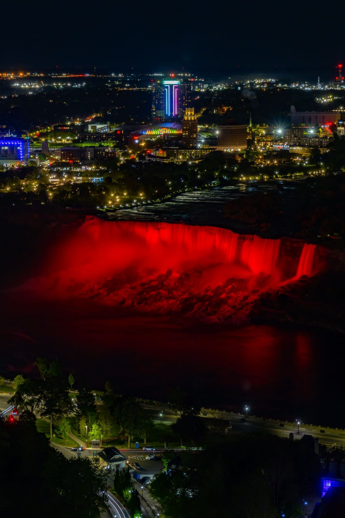 Niagara Falls illuminated in red at night, with city buildings and lights visible in the background.