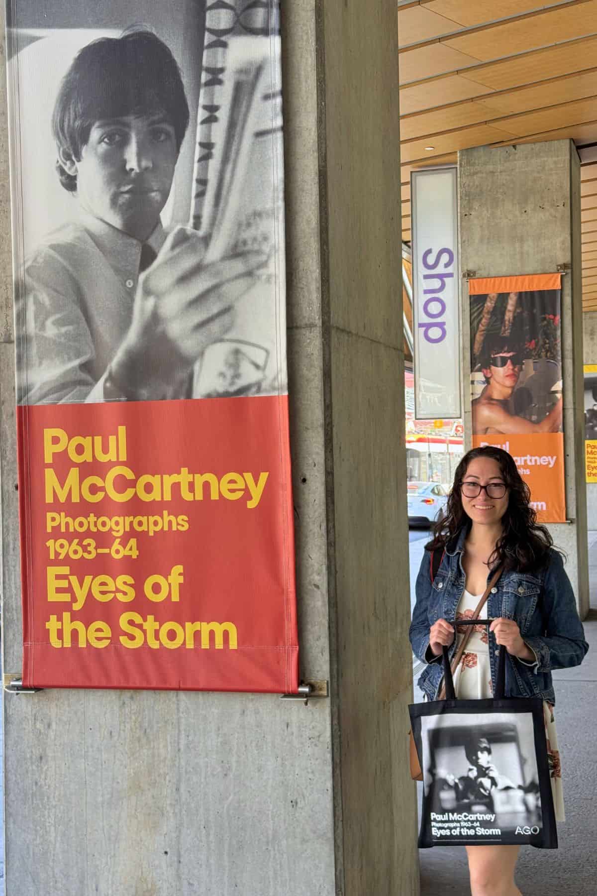 A woman stands next to a “Paul McCartney Photographs 1963-64 Eyes of the Storm” exhibition banner, holding a matching tote bag with the same image and text.