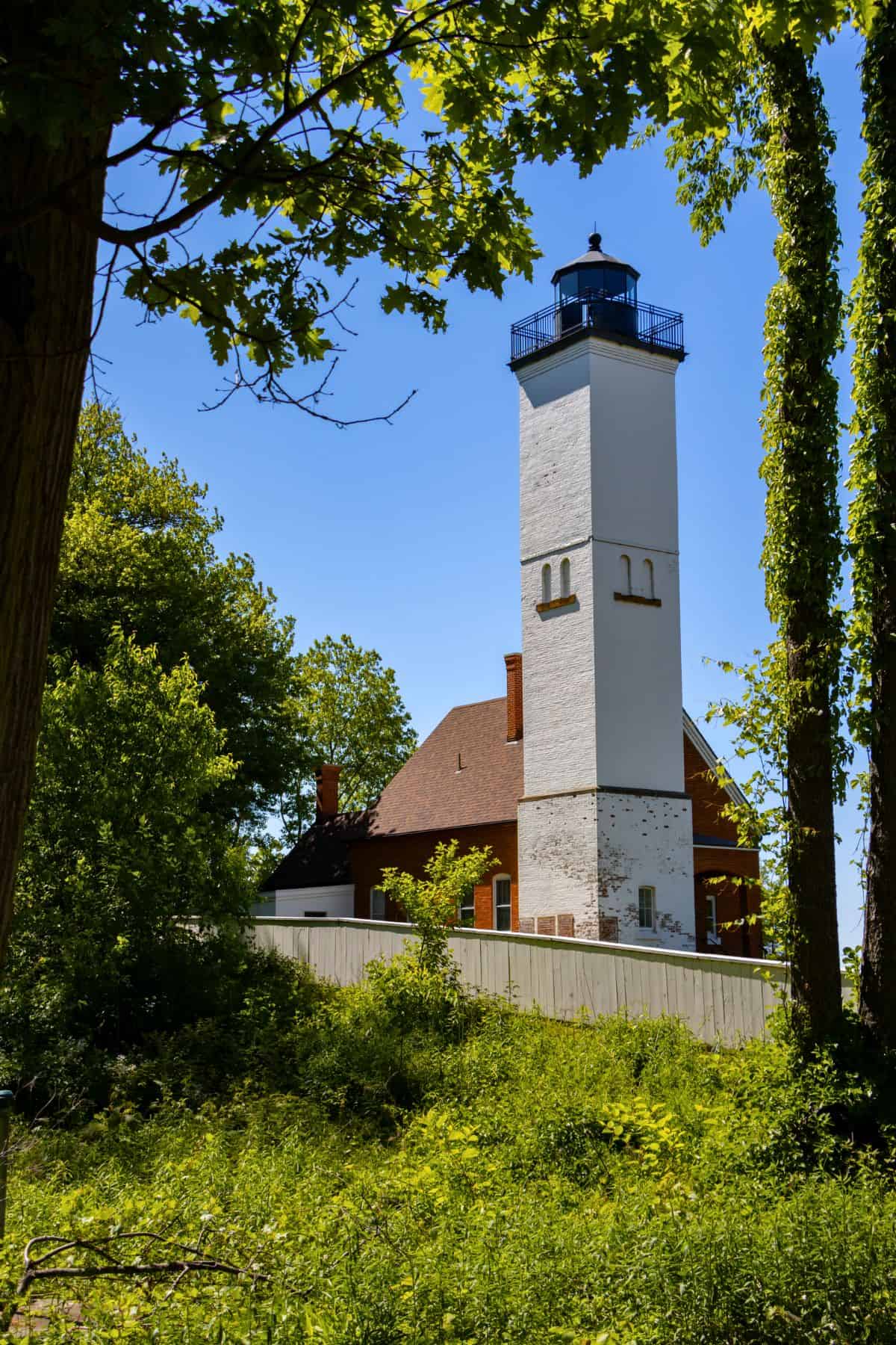 A tall white lighthouse with a black lantern stands next to a brown-roofed building, surrounded by green trees and foliage under a clear blue sky.