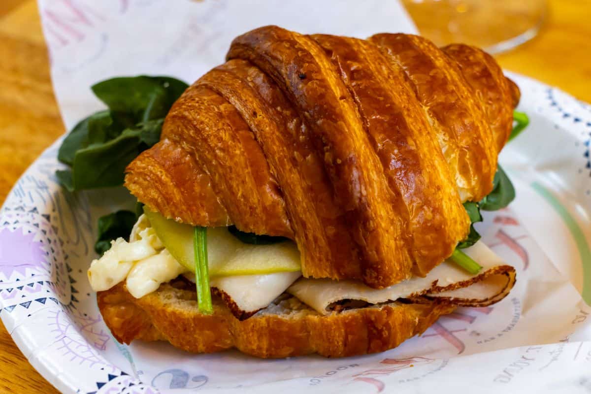 A croissant sandwich filled with turkey, sliced cheese, green apple, and spinach on a paper plate.