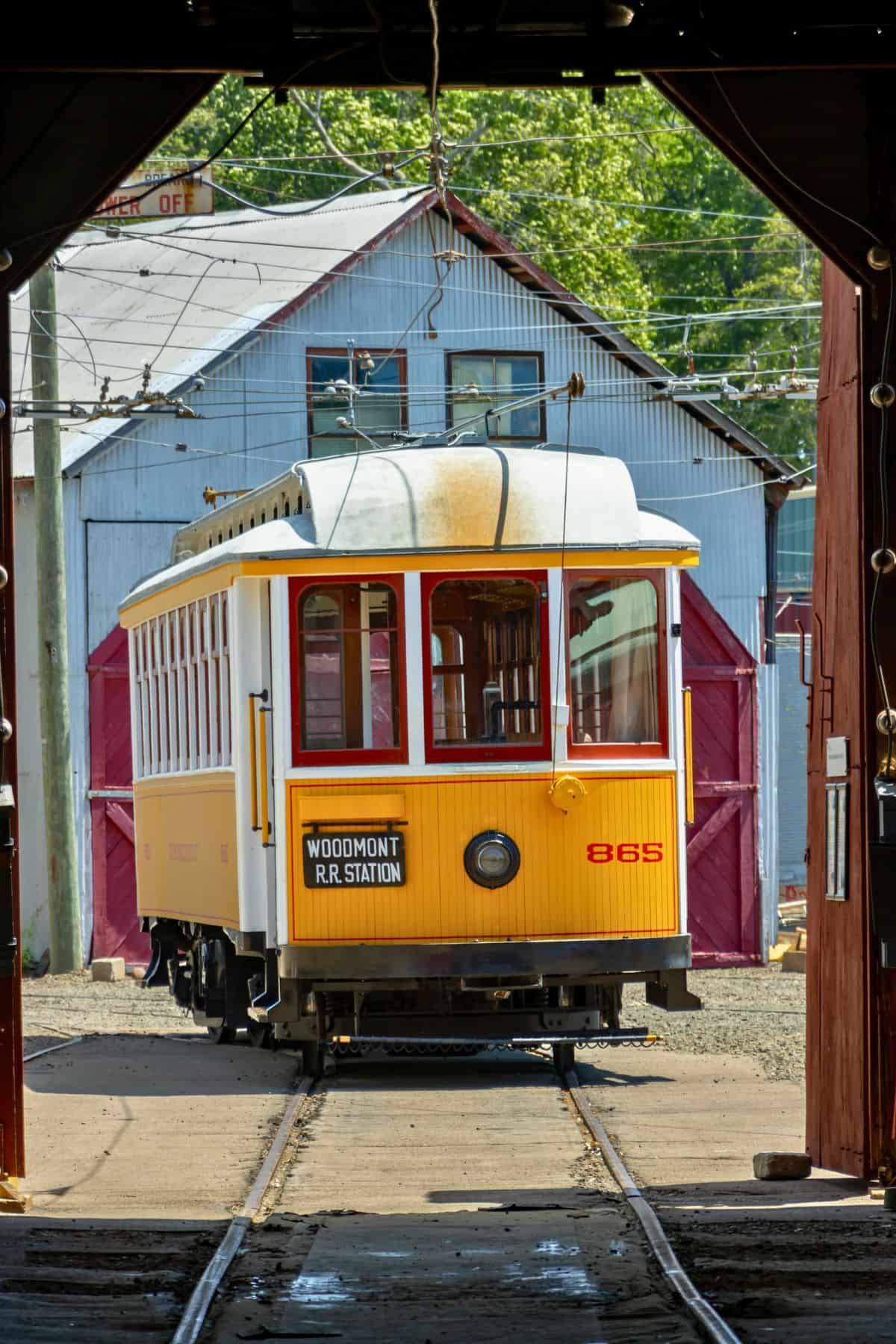 A yellow and white vintage streetcar labeled "Woodmont R.R. Station" sits on tracks outside a red and white barn-style building.