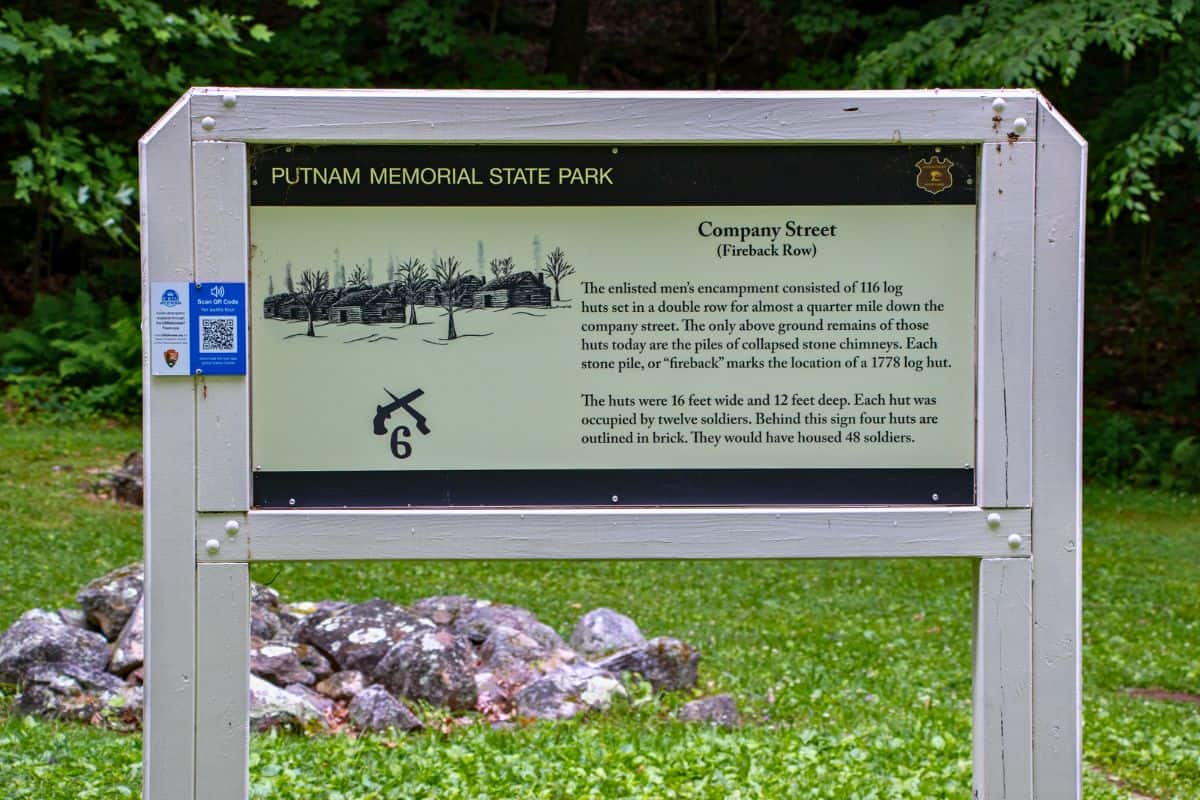 Informational sign at Putnam Memorial State Park describes "Company Street," the site of collapsed log huts from a former military encampment, with a background of trees and rocks.