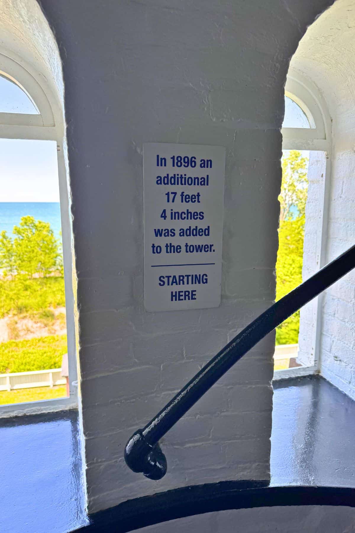 A sign inside a tower reads, “In 1896 an additional 17 feet 4 inches was added to the tower. STARTING HERE,” with windows and a railing visible.