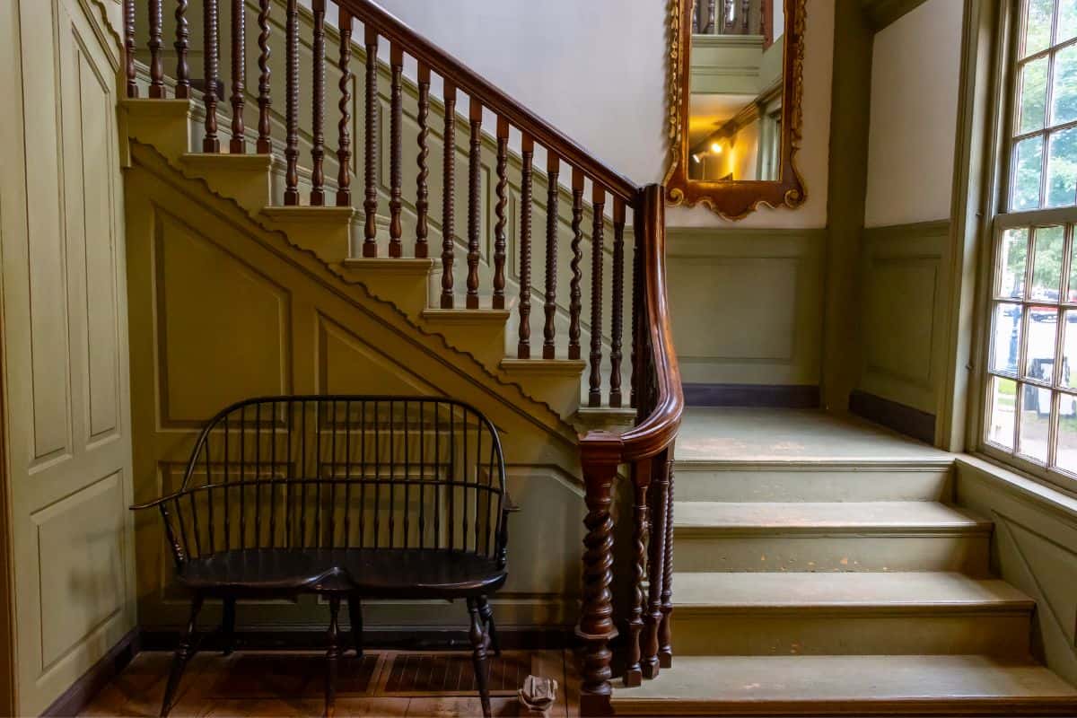A wooden staircase with painted green steps, a large wall mirror, a black wooden bench, and tall windows letting in natural light.