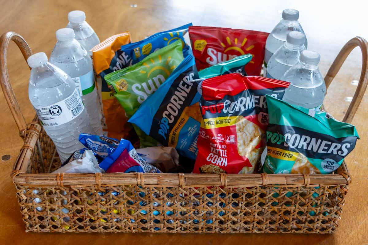 A woven basket holds bottles of water and assorted bags of chips, including PopCorners and SunChips, on a wooden table.