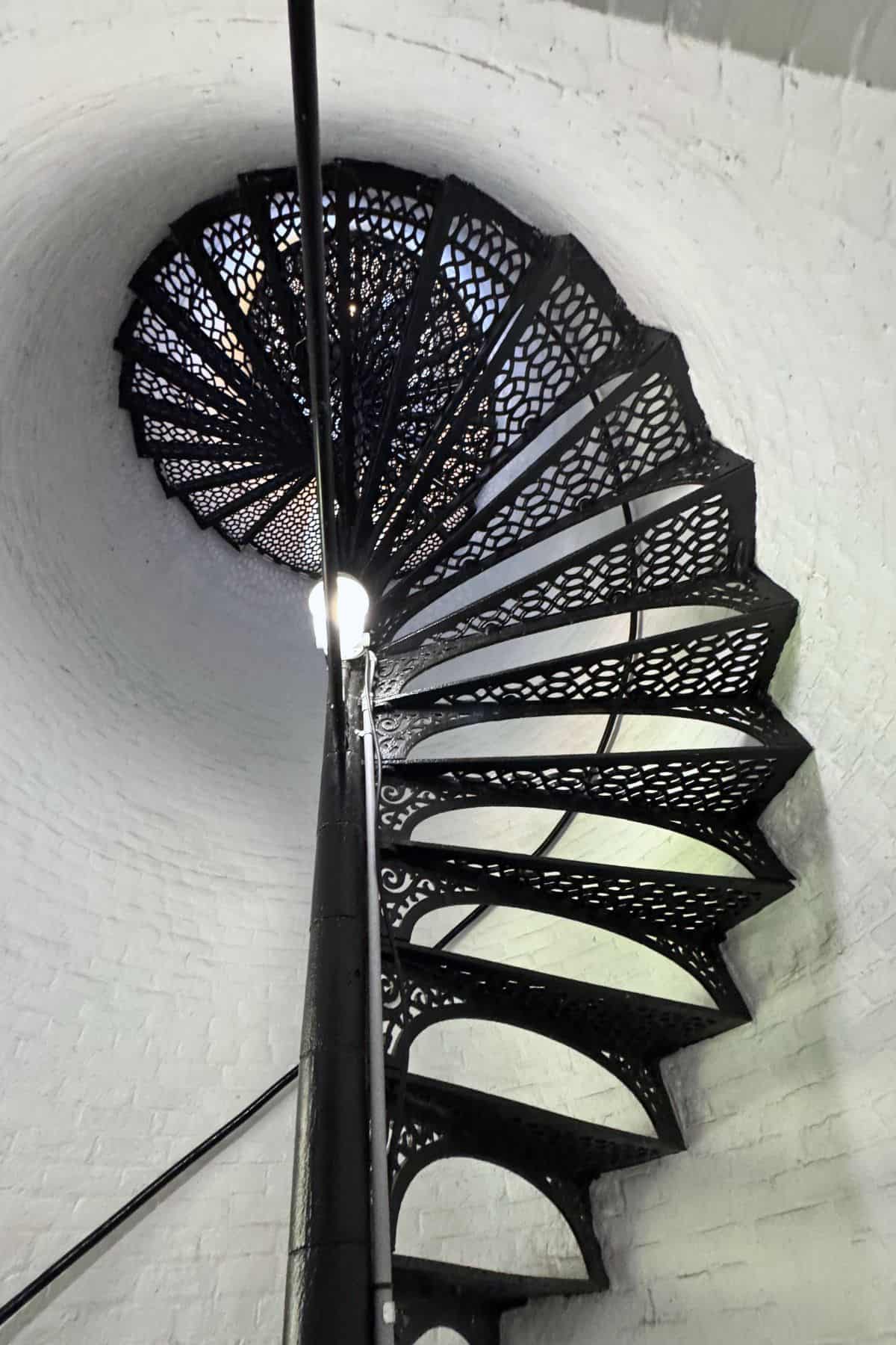 Black wrought iron spiral staircase ascends inside a white brick tower, viewed from the bottom looking upward.
