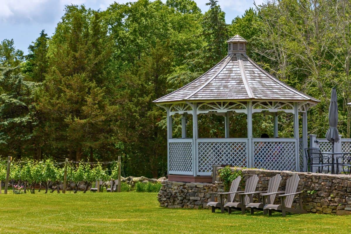 A wooden gazebo stands near Adirondack chairs and a stone wall on a grassy lawn, with trees and grapevines in the background.