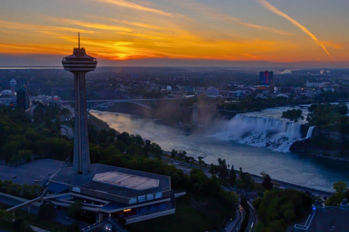 A tall observation tower overlooks Niagara Falls and a river at sunset, with city buildings, a bridge, and colorful sky in the background.