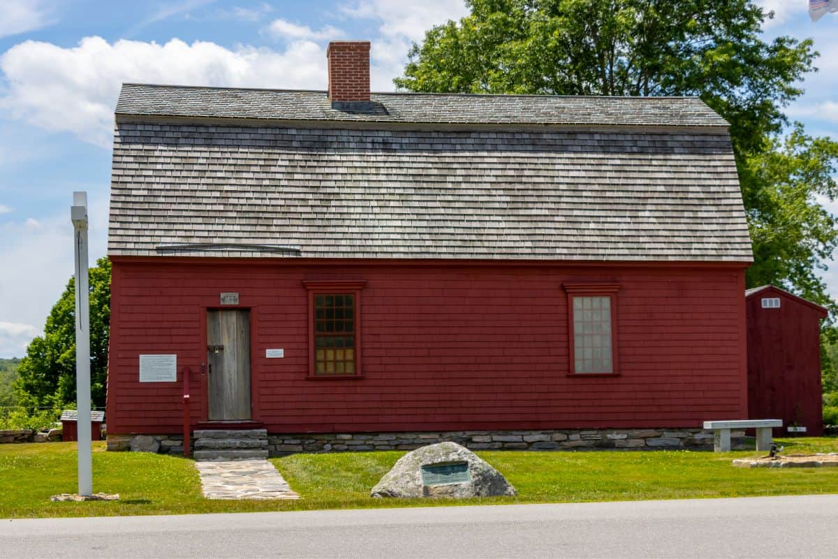 A small, red wooden house with a shingled roof and stone chimney stands on a grassy lawn with a rock monument and a bench out front.
