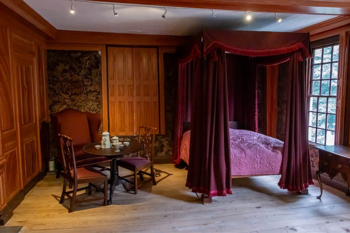 A historic bedroom with wood-paneled walls, a canopy bed with red curtains, a small table set for tea, and two chairs near a window.