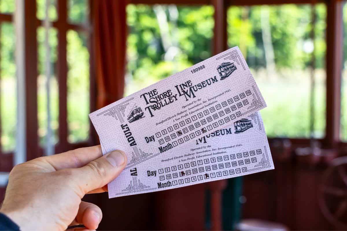A hand holding two tickets for The Shore Line Trolley Museum, with a blurred background of a trolley and greenery outside.