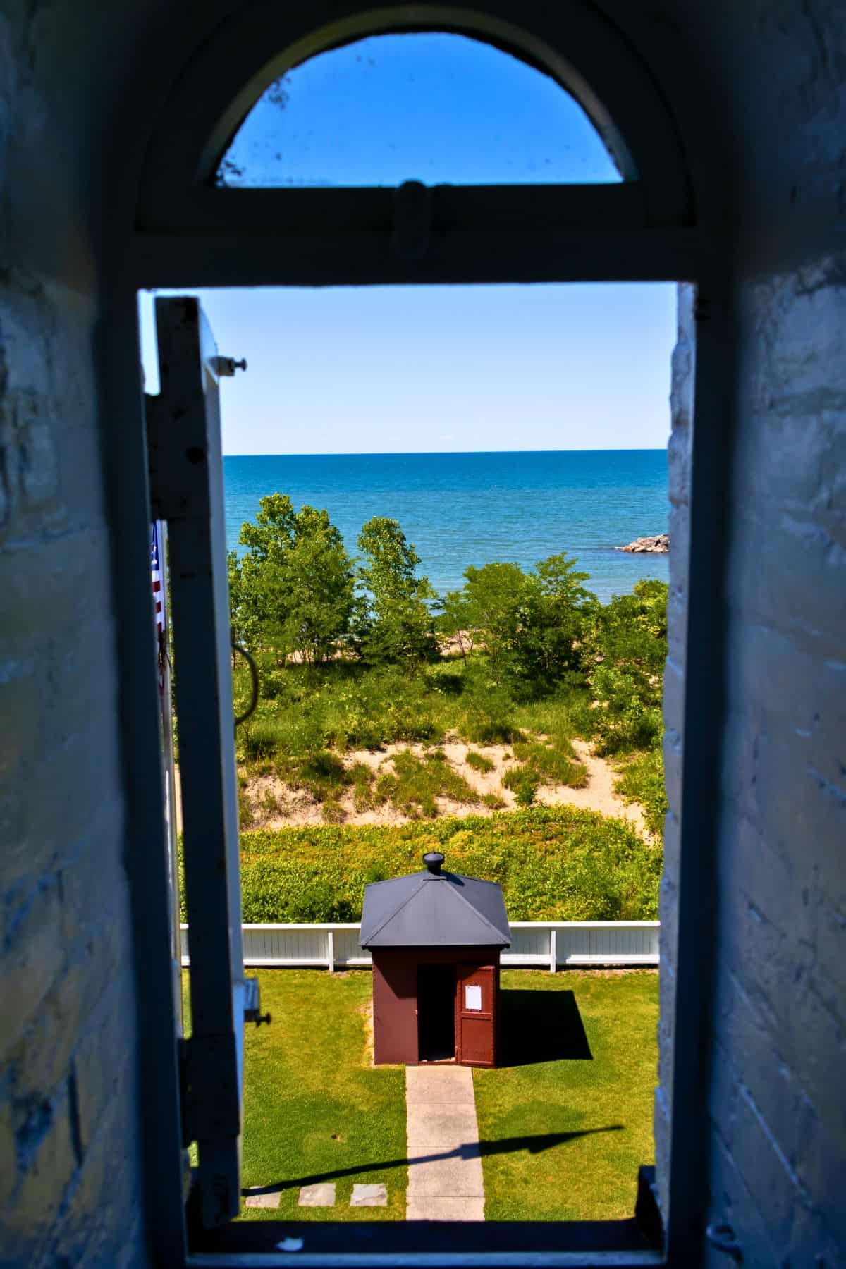 View through an arched window showing a small shed, a path, green trees, and a blue lake under a clear sky.