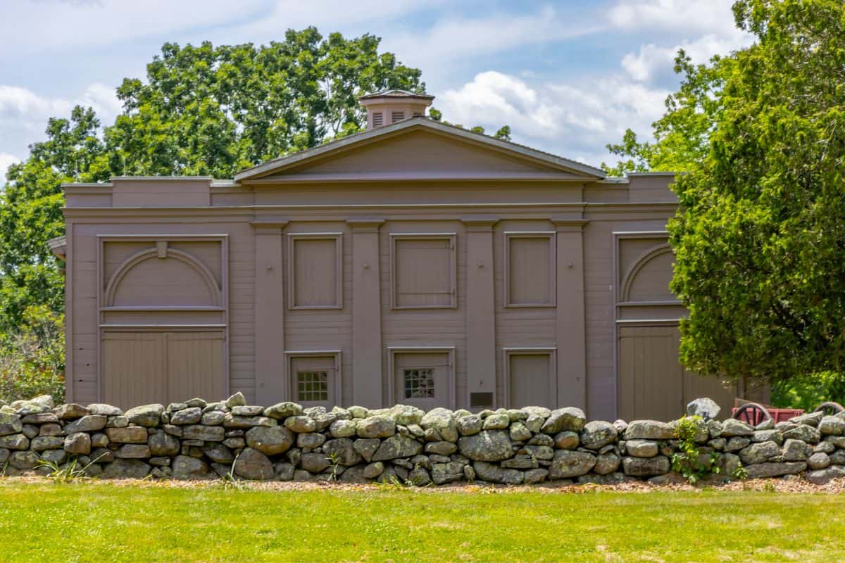 A gray wooden building with closed shutters stands behind a stone wall and trees under a partly cloudy sky.