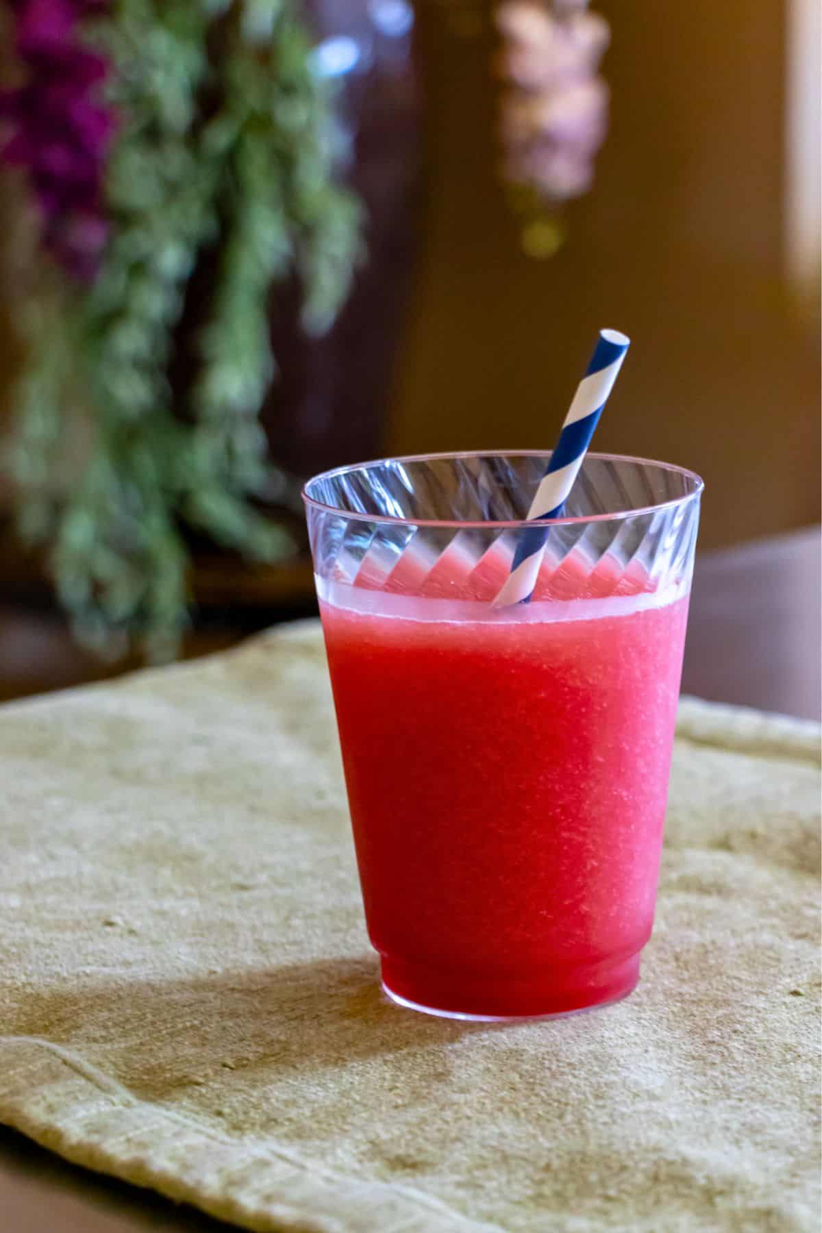 A clear plastic cup filled with a red slushy drink and a blue and white striped straw sits on a beige placemat.
