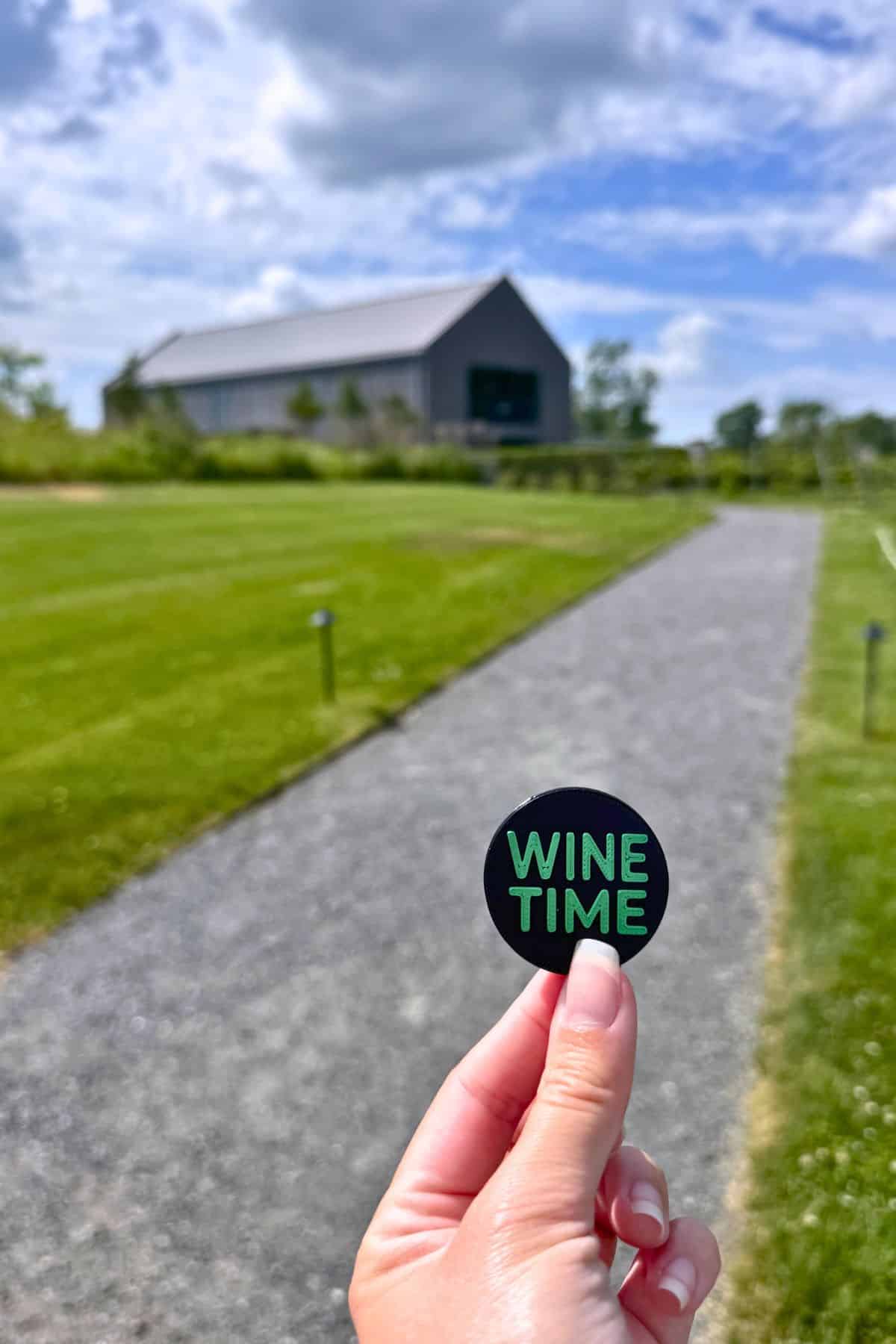 A hand holds a round pin that says "WINE TIME" on a gravel path, with green grass and a barn-like building in the background under a partly cloudy sky.