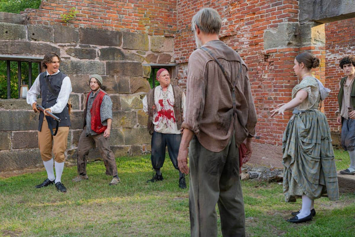 Six people in period costumes stand outside near a brick wall, appearing to be in the middle of an intense conversation or confrontation.