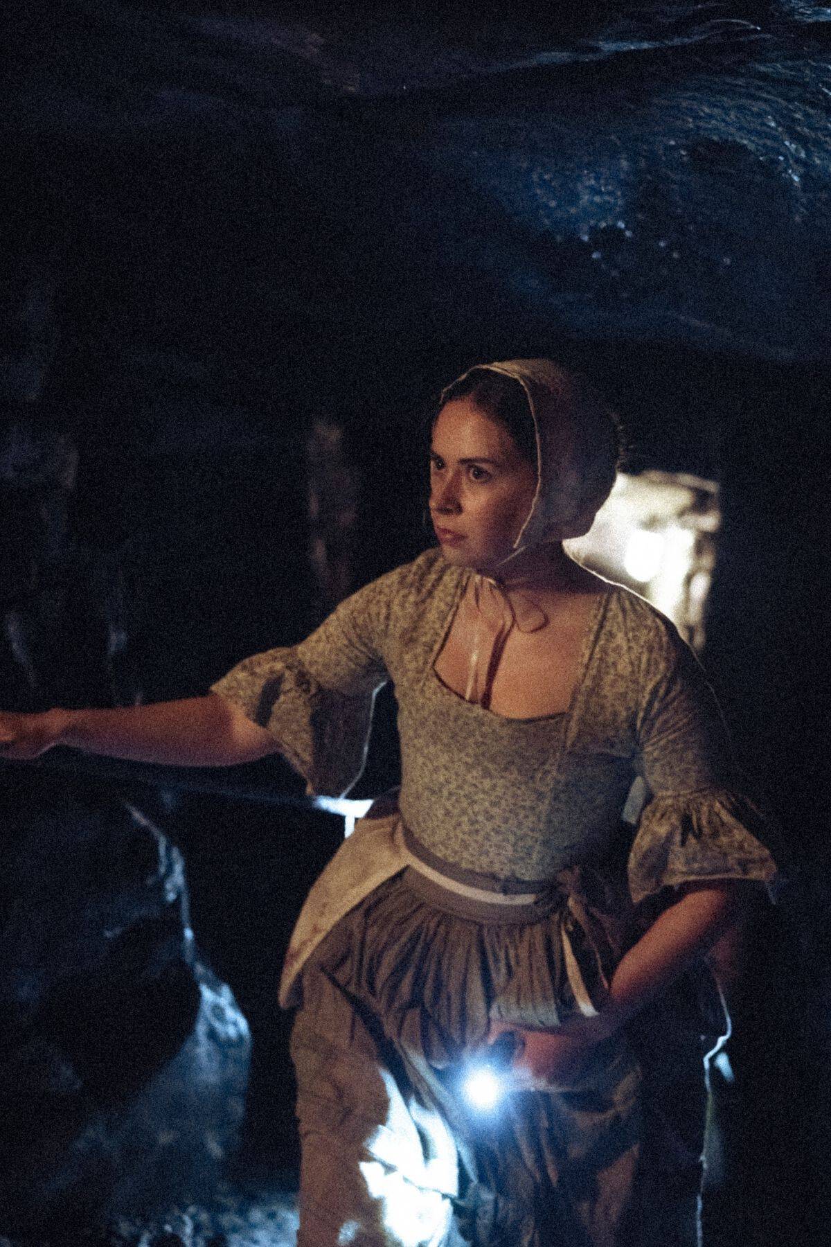 A woman in historical clothing stands in a dimly lit cave, holding a flashlight and looking ahead with a serious expression.