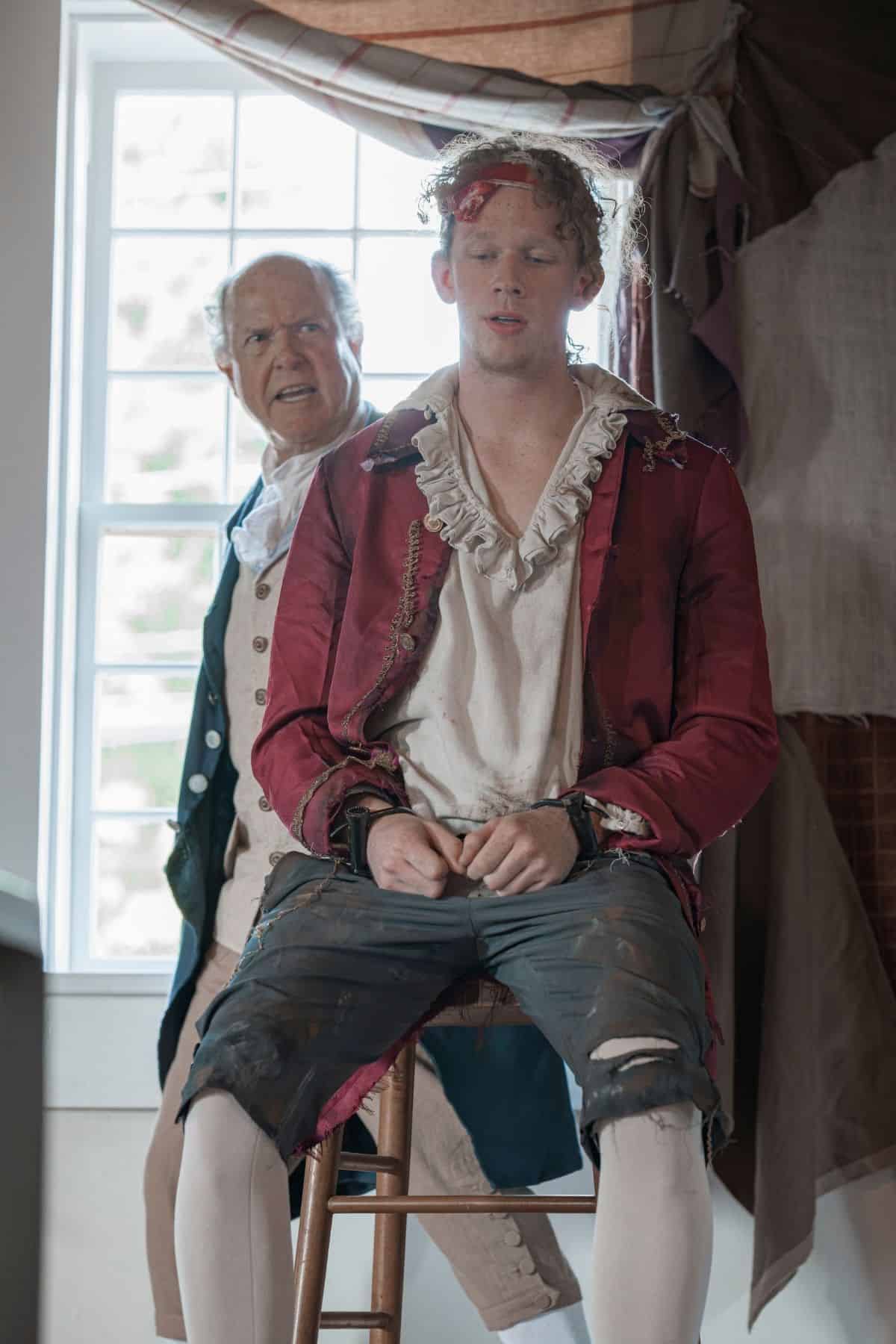 Two men in historical clothing; one sits on a stool with a bloodied forehead and worn clothes, while the other stands behind him, appearing concerned. They are in a room with a window.