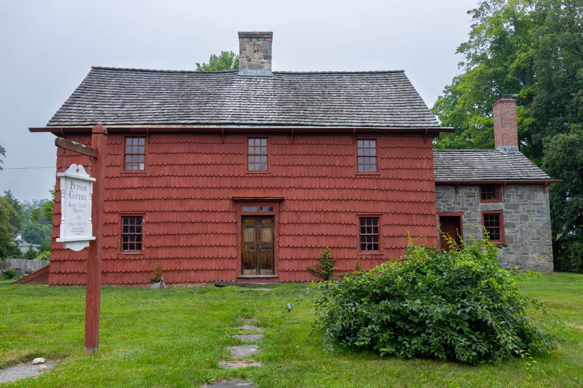 A two-story red colonial house with a shingled roof, stone chimney, and adjacent stone extension, surrounded by grass and trees. A wooden sign stands at the left front.
