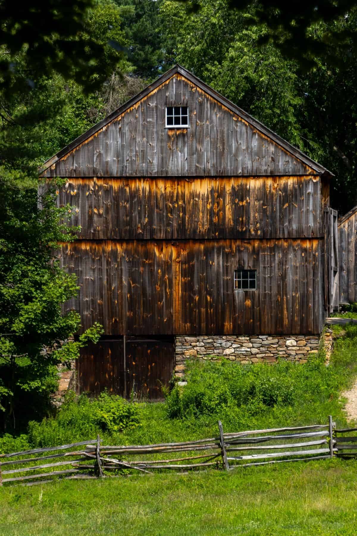 A weathered wooden barn with a stone foundation stands behind a rustic wooden fence, surrounded by green grass and trees on a sunny day.