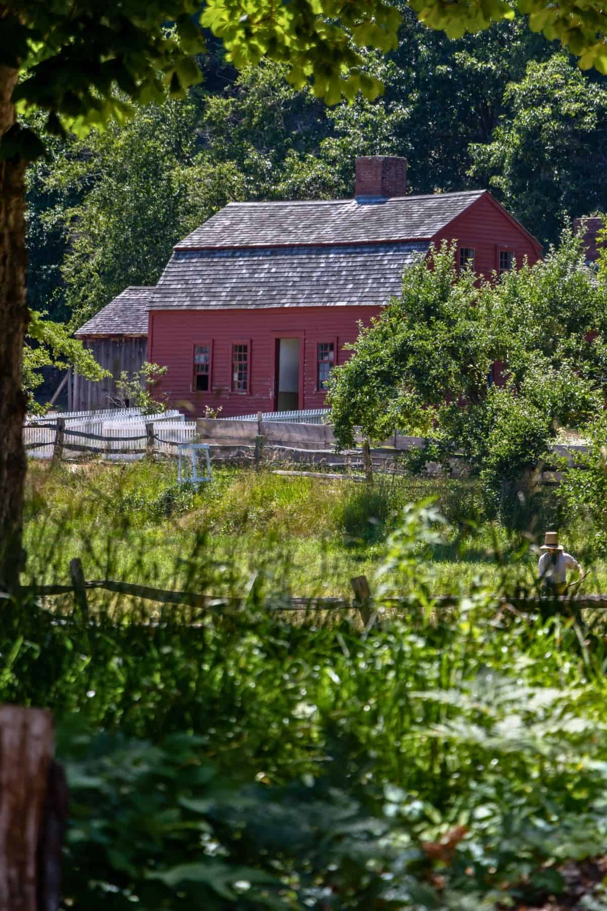 Red wooden house with a shingled roof sits behind a white picket fence, surrounded by trees and greenery on a sunny day.