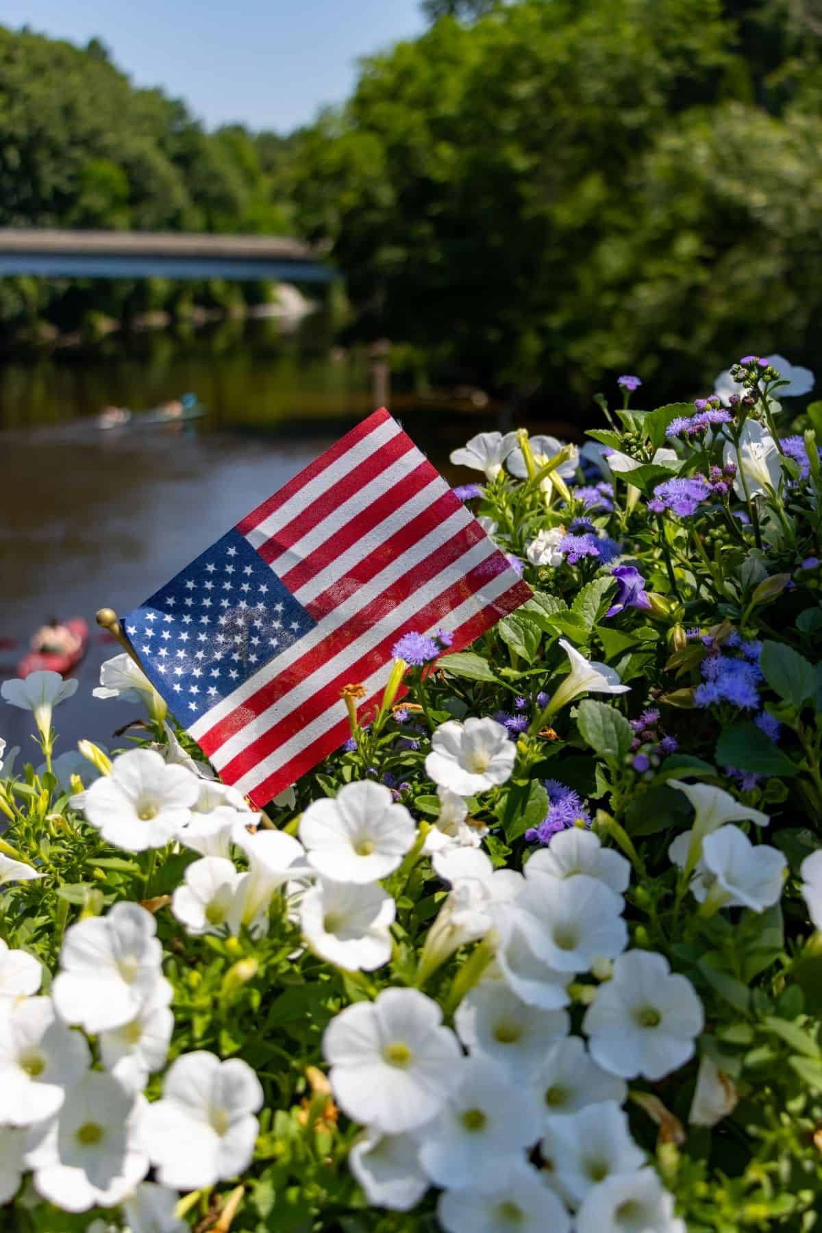 An American flag is displayed among white and purple flowers with a river and bridge in the background on a sunny day.