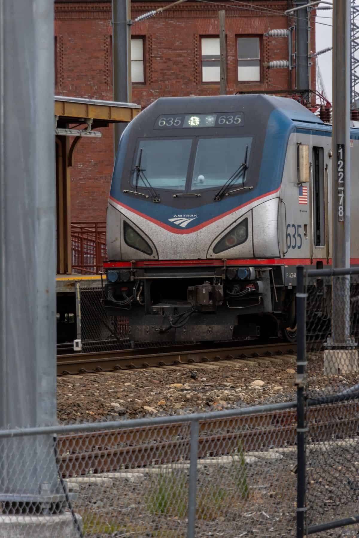 An Amtrak train engine, numbered 635, is stopped on train tracks in front of a brick building, partially obscured by a metal fence and poles.