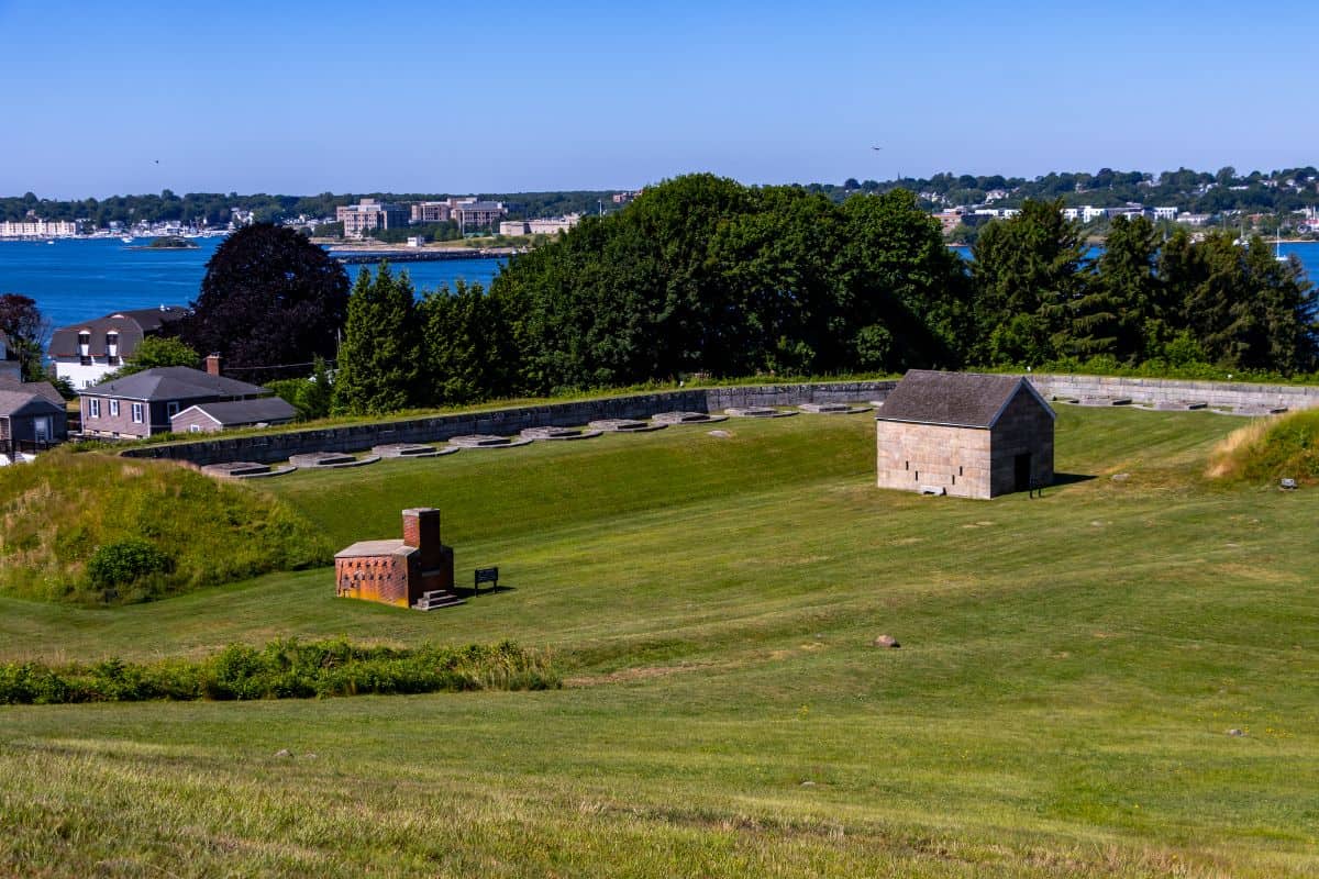 A grassy field with a small stone building and a brick structure, bordered by trees and a stone wall, overlooks water with buildings on the opposite shore.