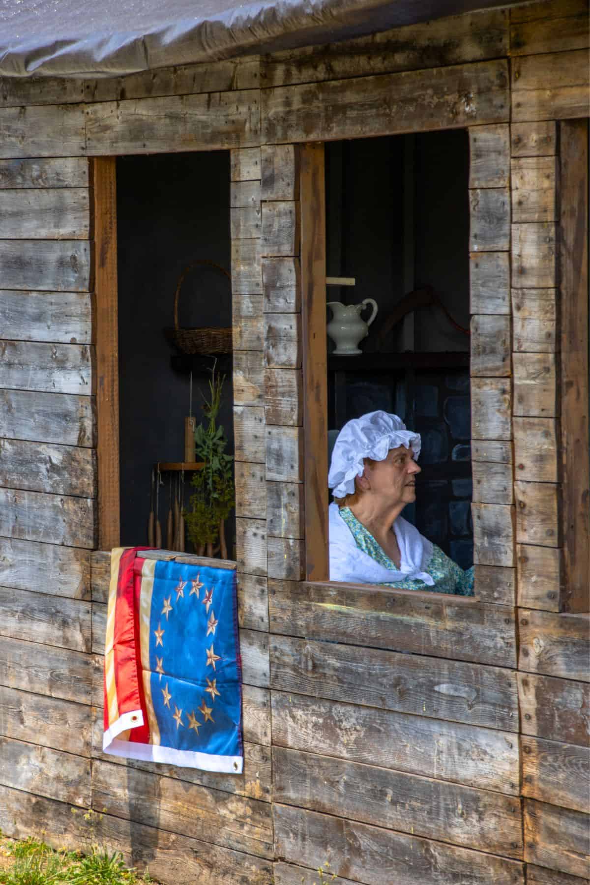 An older woman in a white bonnet sits by an open window in a wooden building. A flag with a circle of stars is draped outside the window.