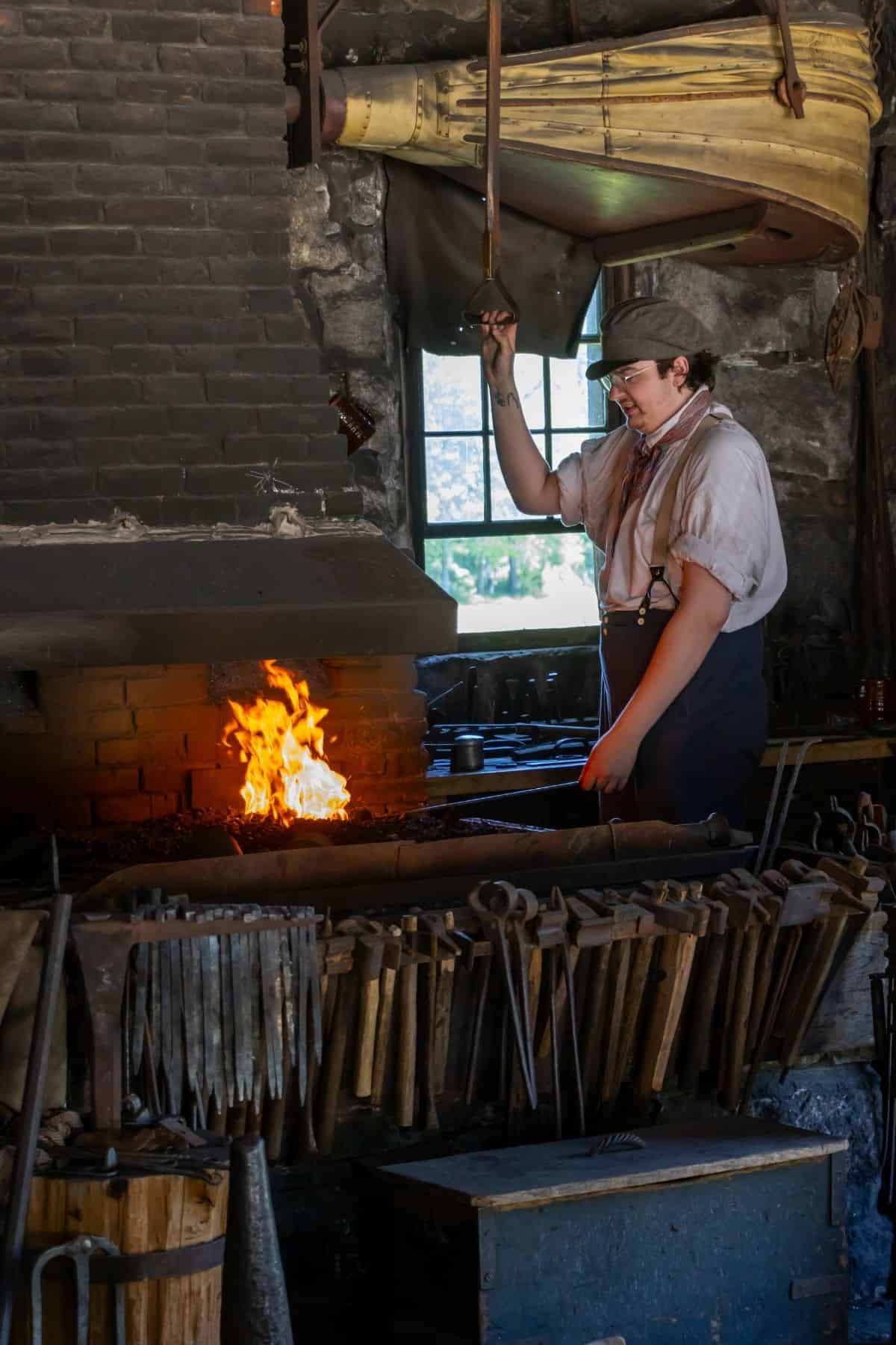 A blacksmith stands by a forge, heating metal over an open flame in a workshop filled with tools hanging on a rack.