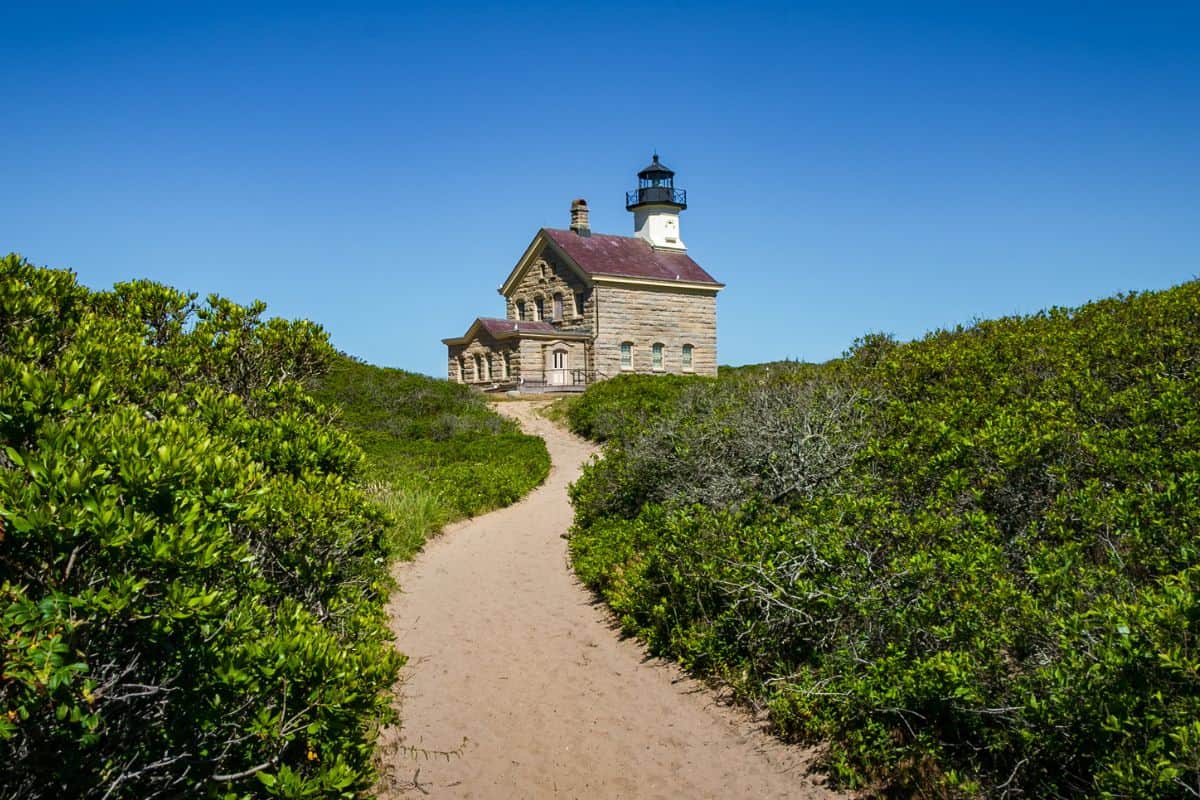 A sandy path lined with green bushes leads to a stone lighthouse under a clear blue sky.