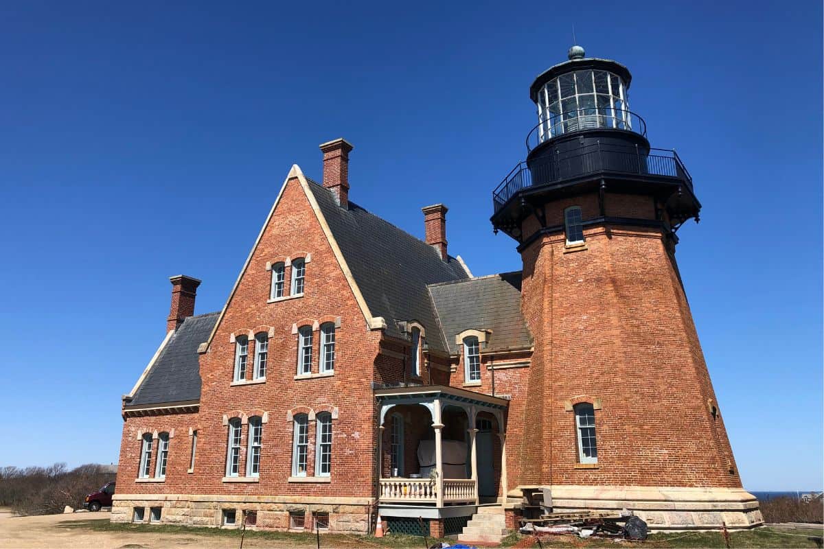 A historic red brick lighthouse with an attached building, set against a clear blue sky on a grassy landscape.