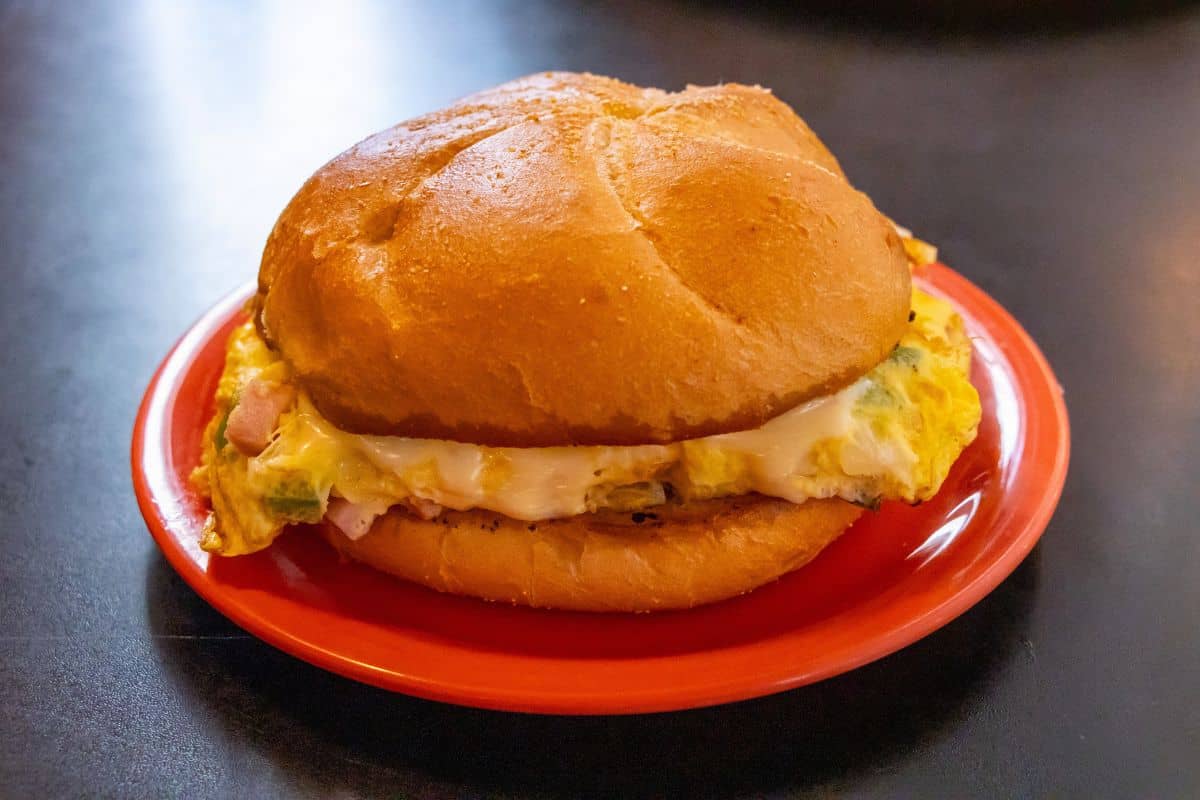 A sandwich with scrambled eggs, melted cheese, and ham is served in a round bun on an orange plate.