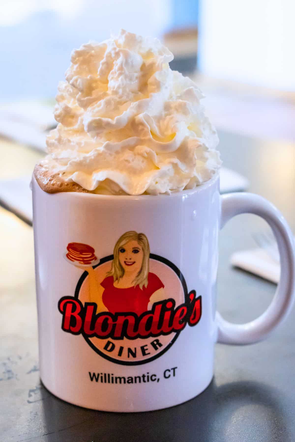 A white mug with the Blondie’s Diner logo, filled with a beverage topped with a large swirl of whipped cream, sits on a table.