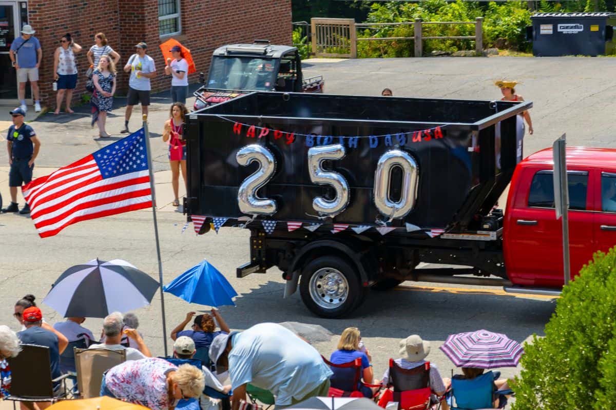 A parade float with metallic "250" balloons and "Happy Birthday USA" sign on a black truck bed; crowds with umbrellas and American flags watch from the roadside.