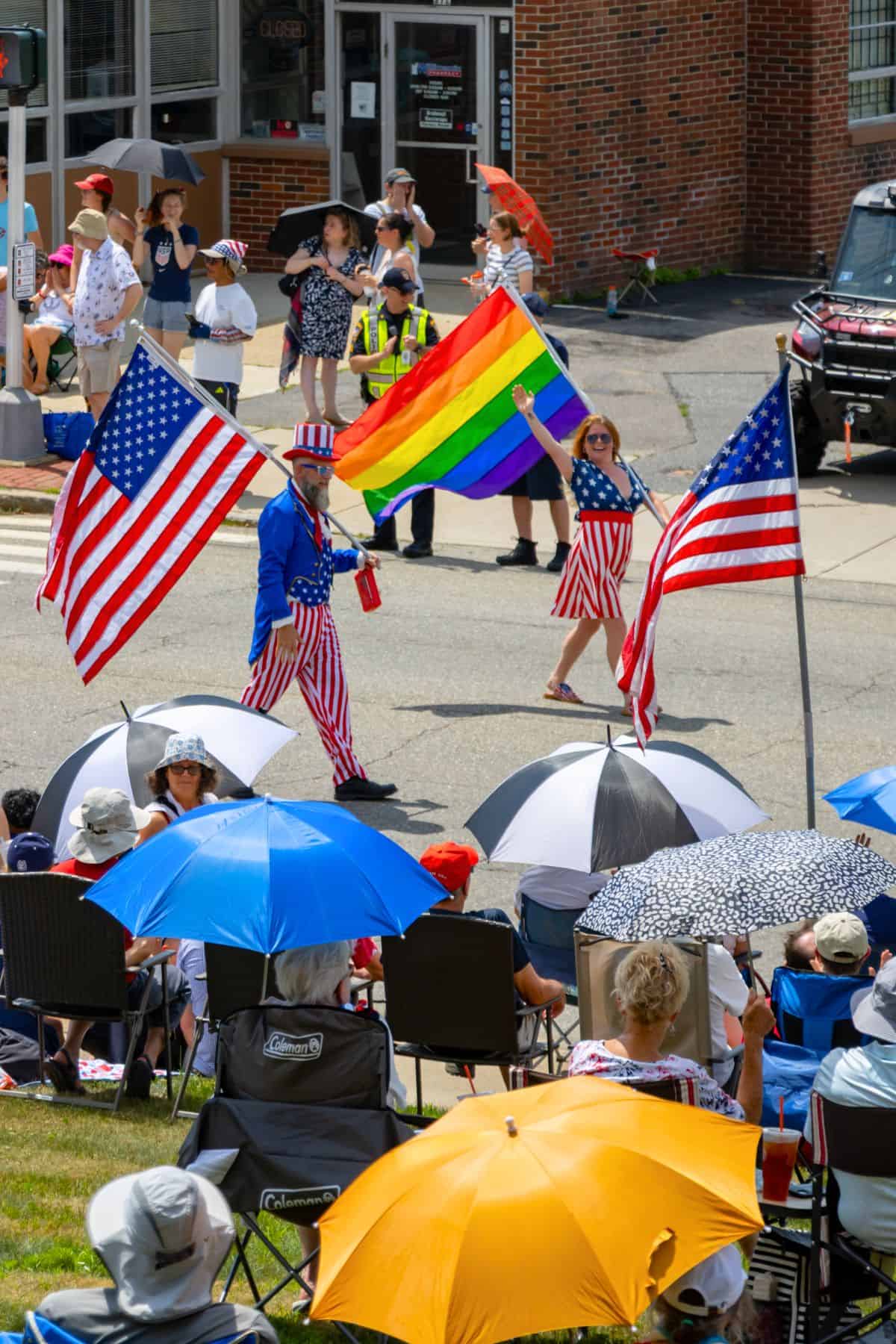 People march in a parade holding two American flags and a rainbow pride flag, while spectators watch from under umbrellas on the sidewalk.
