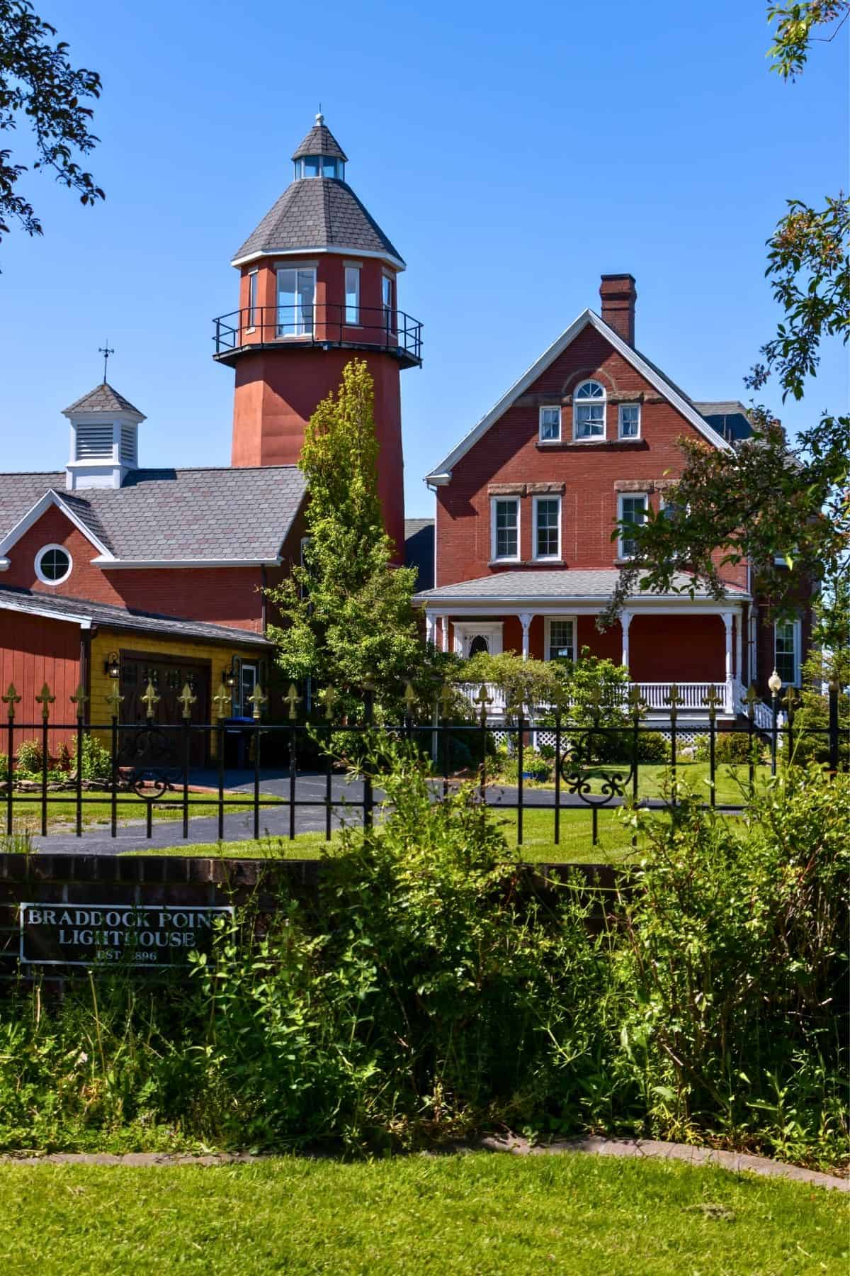 Red brick lighthouse with attached house behind a black iron fence and a sign reading "Braddock Point Lighthouse" on a sunny day with clear blue sky.