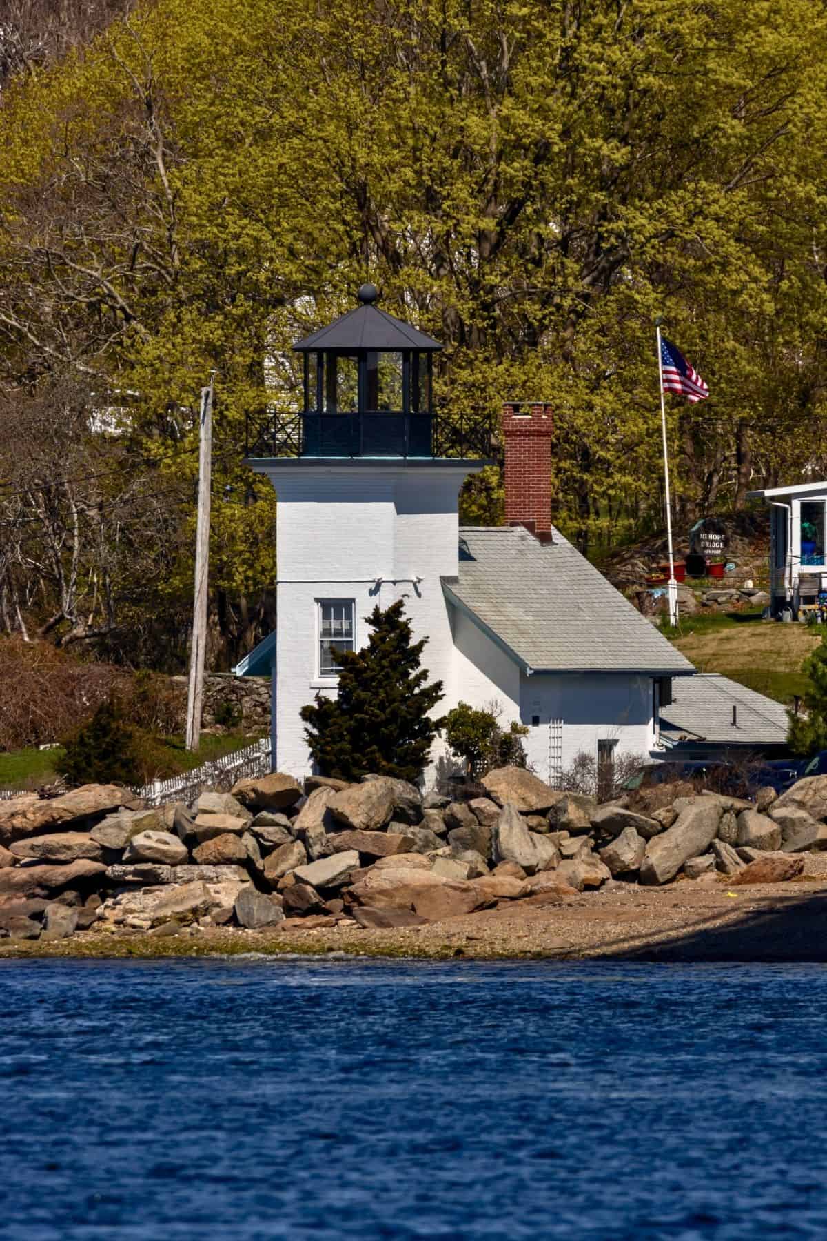 A small white lighthouse with a black lantern sits by rocky shore, surrounded by trees and a U.S. flag, near calm blue water.