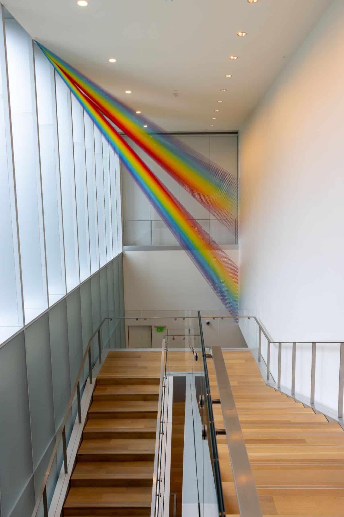 Colorful thread art installation forms a rainbow diagonally across a modern stairwell with wooden steps, glass railings, and white walls.