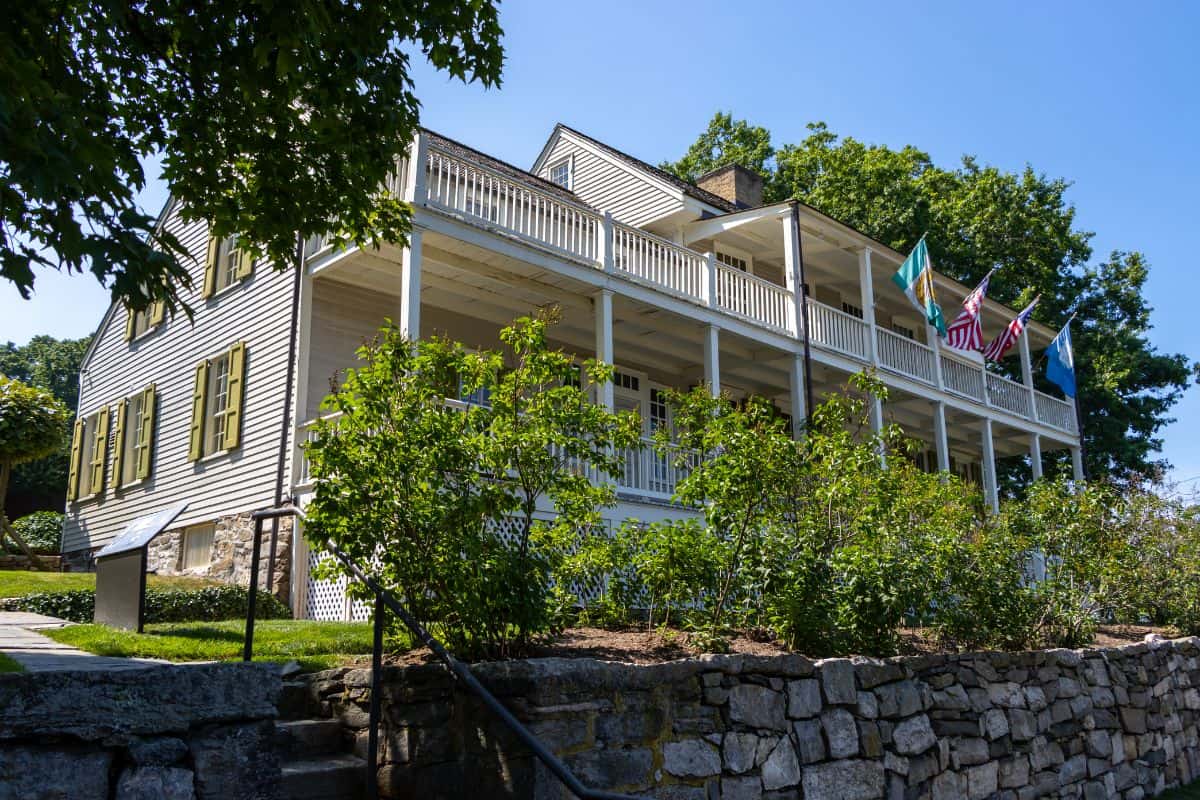 A large two-story white house with green shutters, wraparound porch, and multiple flags, surrounded by trees and a stone wall.