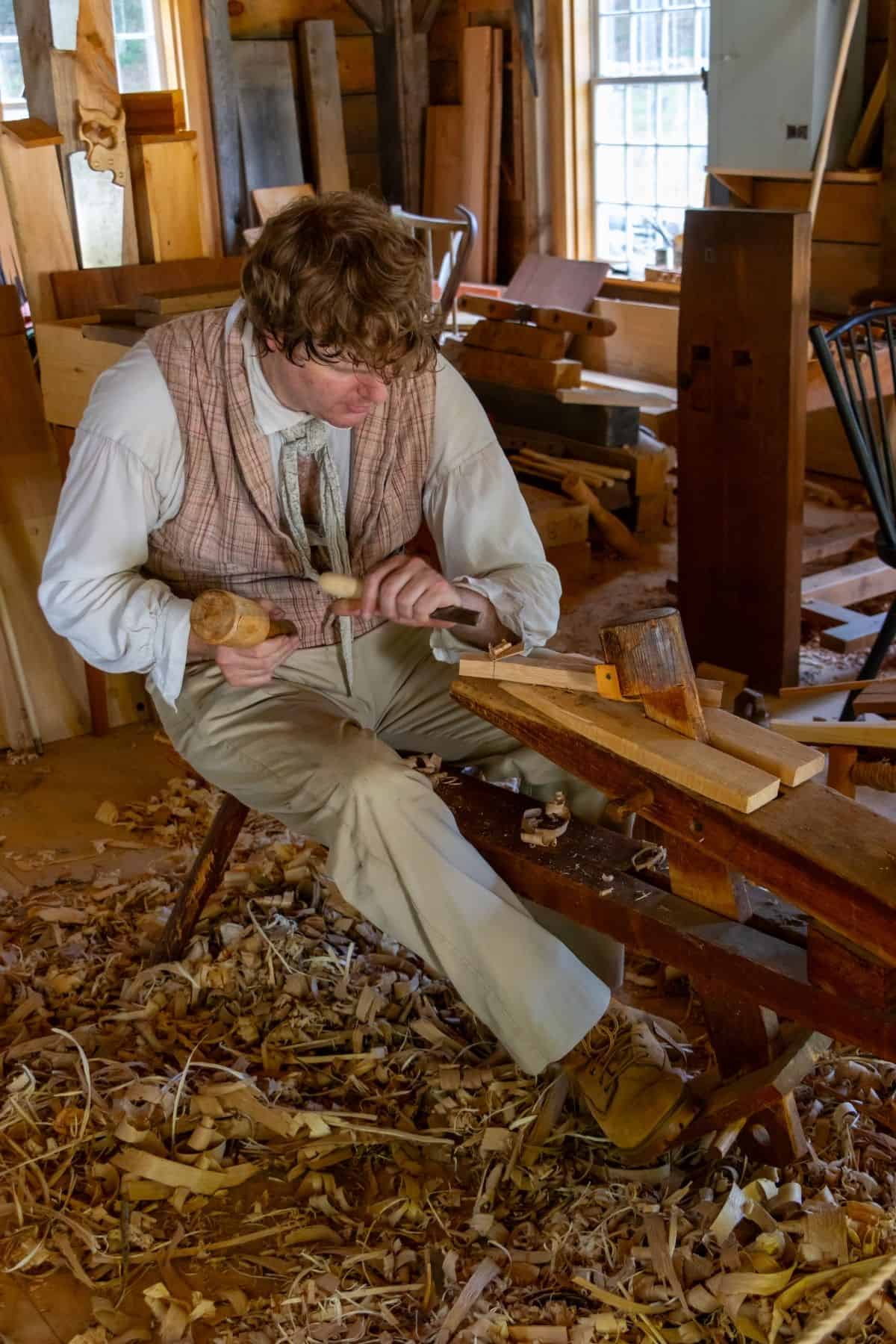 A man in historical clothing carves wood with a chisel and mallet in a workshop filled with wood shavings and woodworking tools.