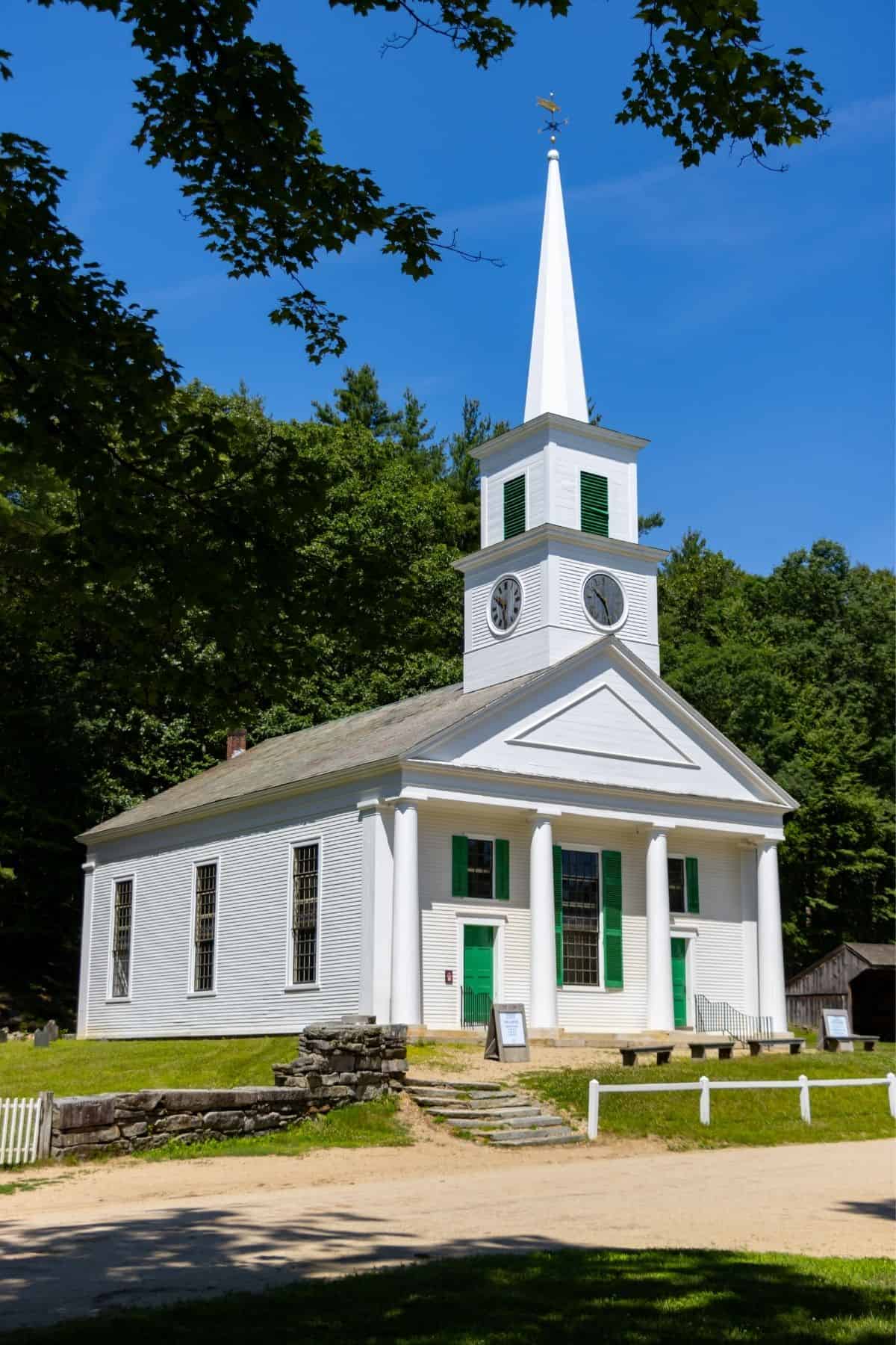 White church with green doors, tall steeple, and clock tower, set against a backdrop of trees under a clear blue sky.